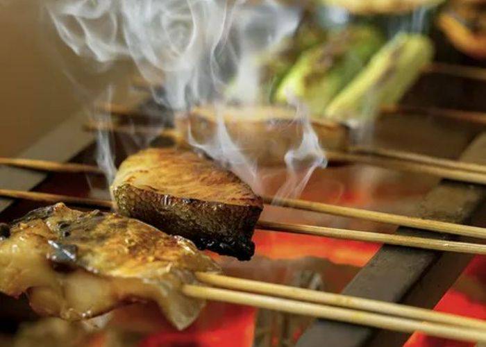 Skewered fish being grilled; smoke is rising from the fish as it crisps up.